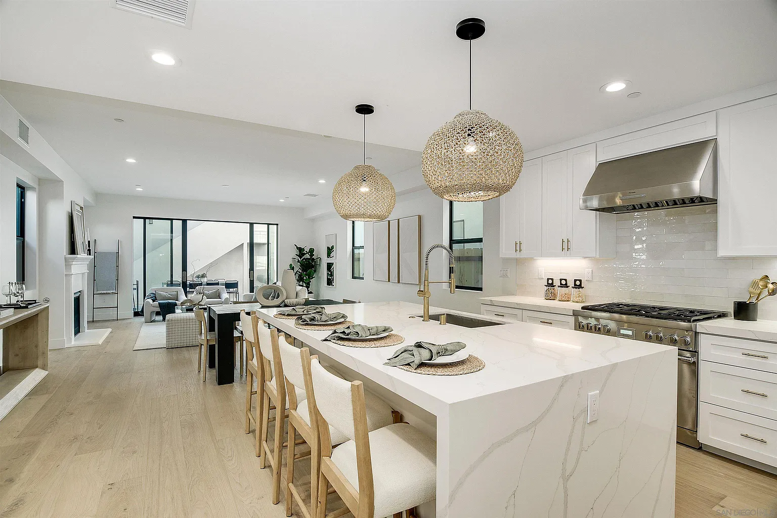 Modern kitchen with a large marble island, four wooden barstools, pendant lights, white cabinets, and stainless steel appliances, opening to a living area with light wood flooring.